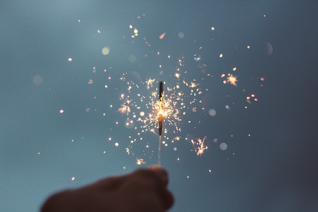 A hand holding a sparkling firework against a dusk blue sky, with bright sparks scattering around