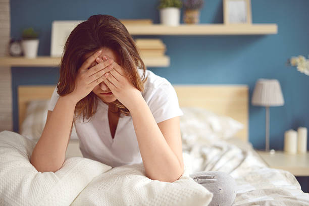A distressed woman sitting on her bed with her face in her hands, appearing to be under stress.