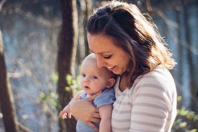 A smiling woman holding an infant in a sunlit natural setting.