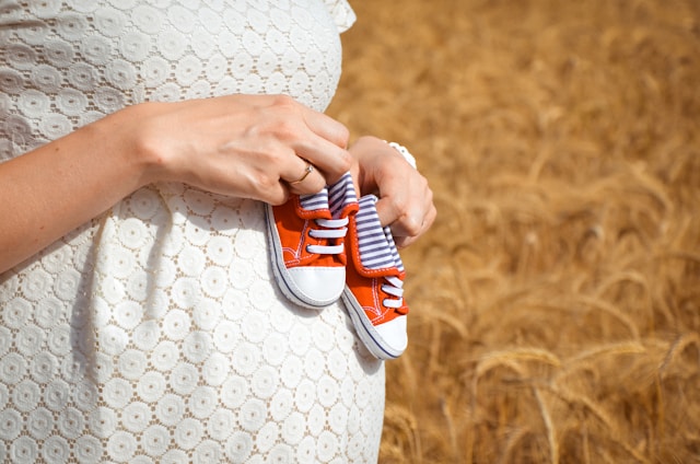 A pregnant woman holding a pair of tiny orange sneakers against her belly in a wheat field to represent miscarriage prevention and keeping a pregnancy till full term