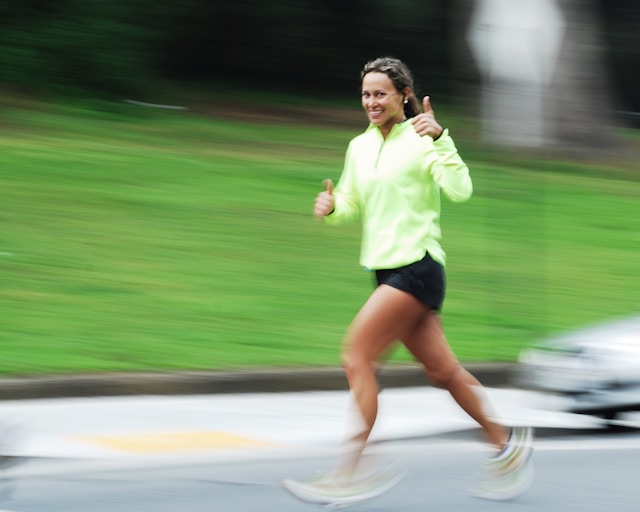 A woman in a neon green jacket gives a thumbs up while jogging in an urban setting.