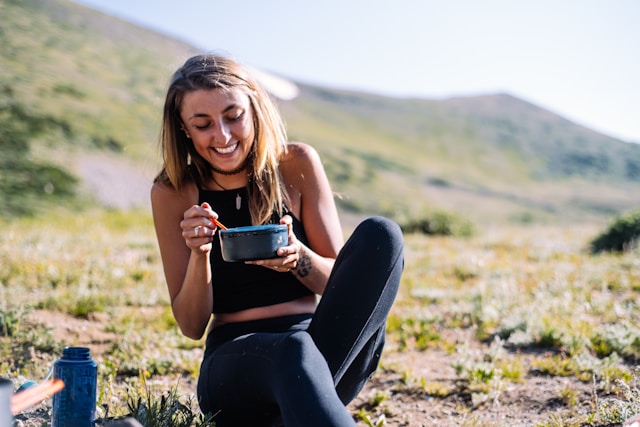 A joyful woman sitting outdoors, eating from a bowl, surrounded by a mountain landscape.