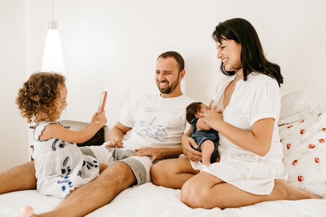 A joyful family on a bed with a toddler holding a pregnancy test, indicating a positive result to the parents.