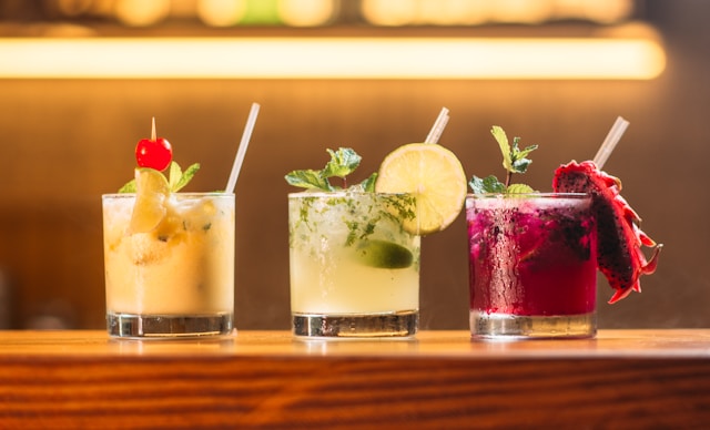 Three colorful cocktails garnished with fruit and herbs lined up on a bar.