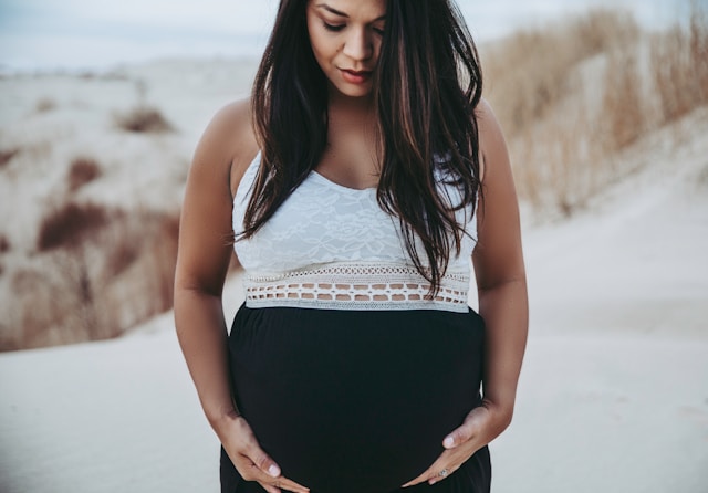 A contemplative pregnant woman standing on a sandy landscape, hands gently supporting her belly.