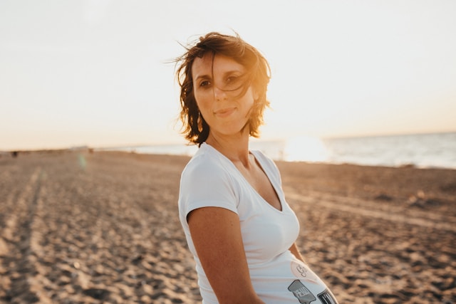 A smiling pregnant woman standing on a beach at sunset, hand on her hip, looking optimistically towards the future.