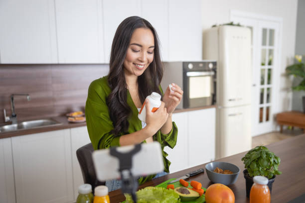 A woman in a green shirt smiles while holding a supplement bottle, with fresh vegetables on the kitchen counter, ready for a healthy meal.
