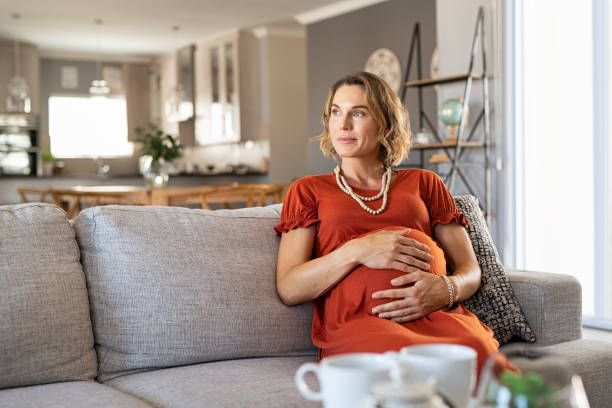 A pregnant woman in a rust-colored dress sits contemplatively on a sofa, her hands gently cradling her belly.