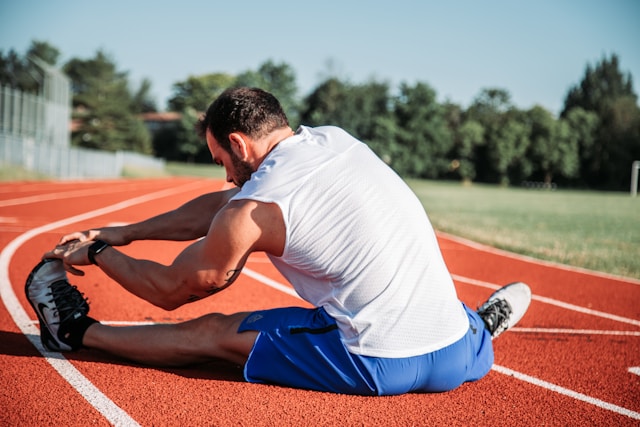 An athletic man stretching on a red running track field, reaching for his toes, dressed in a white tank top and blue shorts.