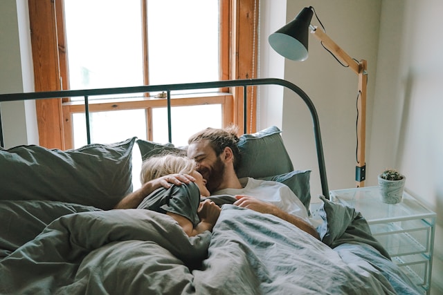 A couple lies in bed together, facing each other and sharing an intimate moment.